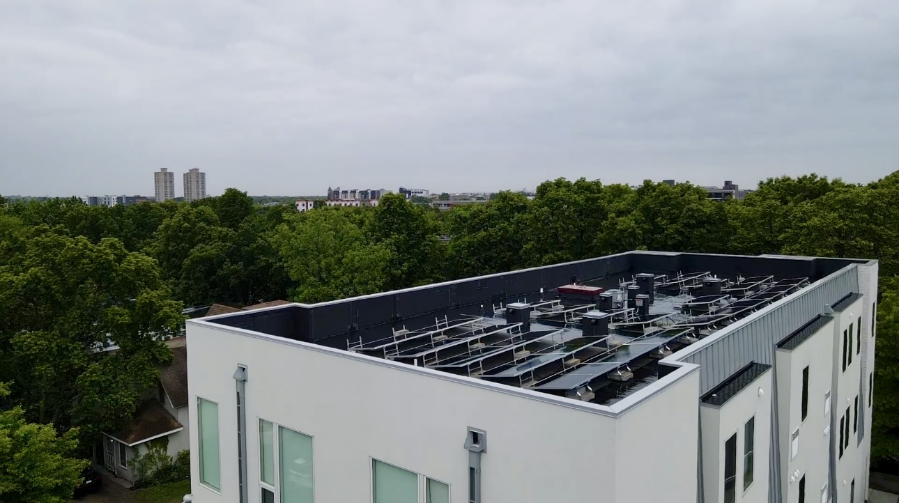Side aerial view of flat roof with solar panels and HVAC, surrounded by trees and mid-rise buildings in Minneapolis.