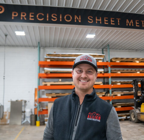 Smiling Allied Construction team member standing inside Precision Sheet Metal facility with safety gear and materials in the background