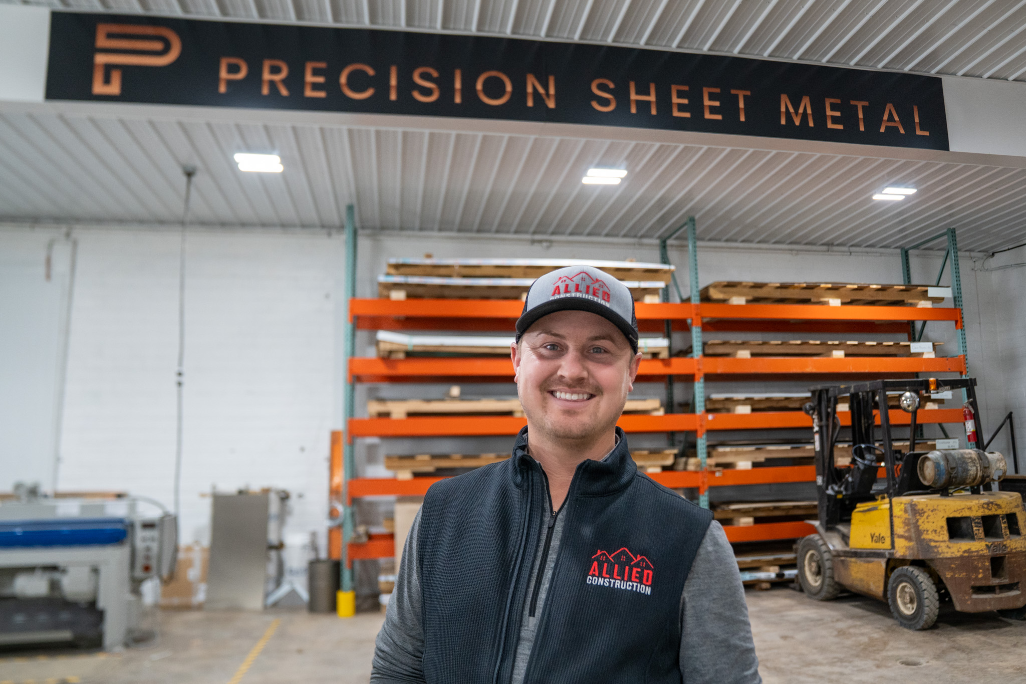 Smiling Allied Construction team member standing inside Precision Sheet Metal facility with safety gear and materials in the background