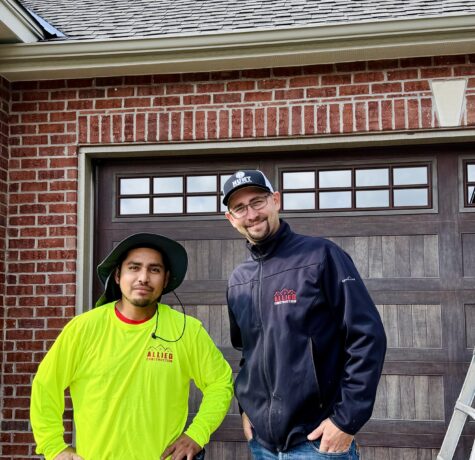Two Allied Construction team members standing in front of a brick home and garage, smiling at the camera.